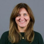 Woman with long brown hair with highlights wearing a dark green sweater and a gold necklace.