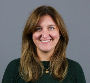 Woman with long brown hair with highlights wearing a dark green sweater and a gold necklace.