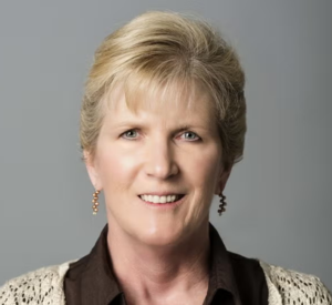 Woman with short blonde hair wearing a white crochet cardigan over a brown collared blouse.