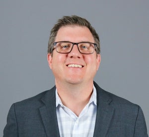 Man with short brown hair wearing glasses and a dark gray blazer over a light gray collared shirt.