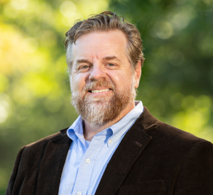 Man with light brown and gray hair and a beard wearing a dark brown suit jacket over a light blue collared shirt.