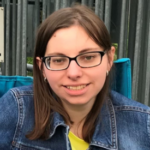 Woman with brown hair and dark glasses wearing a denim jacket over a yellow shirt.