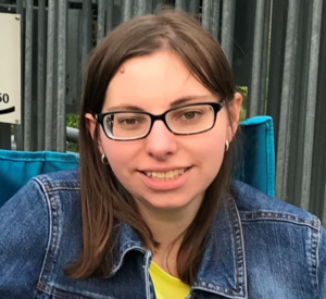 Woman with brown hair and dark glasses wearing a denim jacket over a yellow shirt.