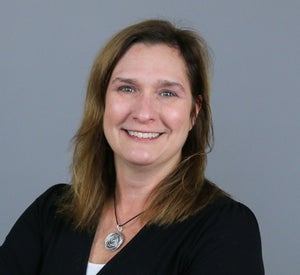 Woman with brown hair wearing a black sweater over a white top and a necklace with a silver pendant.