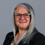 Woman with long gray hair wearing black glasses and a black blazer over a brown striped blouse.