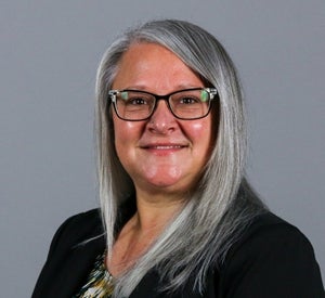 Woman with long gray hair wearing black glasses and a black blazer over a brown striped blouse.