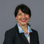 Woman with dark hair pulled back wearing gold earrings and a black blazer over a white collared shirt with white, pink and blue color patterns.