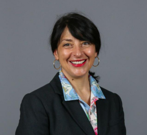 Woman with dark hair pulled back wearing gold earrings and a black blazer over a white collared shirt with white, pink and blue color patterns.