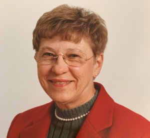 Woman with red hair and wire glasses wearing a red blazer over a dark gray sweater and a white pearl necklace.