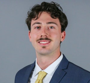 Man with brown curly hair and a mustache wearing a navy blue suit jacket over a white collared shirt and a yellow tie with polka dots.