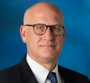 Bald man wearing a black suit jacket over a white collared shirt and a dark blue tie with white dots.