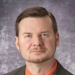 A man with brown hair wearing a brown suit jacket over an orange collared shirt.