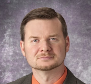 A man with brown hair wearing a brown suit jacket over an orange collared shirt.