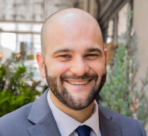 Bald man with a beard wearing a a gray suit jacket over a white collared shirt and a dark blue tie.