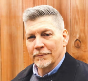 Man with white hair wearing a black jacket over a gray sweater and a blue collared shirt.