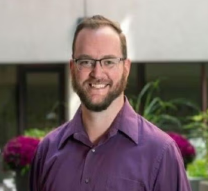 Man with auburn hair and a beard wearing a purple collared shirt.