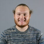 Man with red hair and a beard wearing a blue and tan striped sweater.