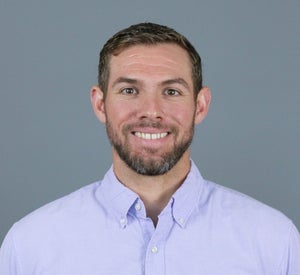 Man with short brown hair and a beard wearing a light purple collared shirt.