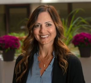 Woman with brown curled hair wearing a black cardigan over a blue blouse.