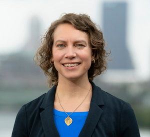 Woman with short brown hair wearing a black blazer over a blue shirt and a gold leaf necklace.