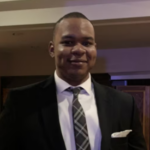 Man with short black hair wearing a black suit jacket over a white collared shirt and a black and white plaid tie.