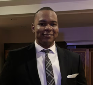 Man with short black hair wearing a black suit jacket over a white collared shirt and a black and white plaid tie.