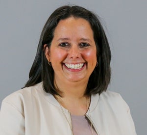 Woman with short brown hair wearing a tan blazer over a light pink top and a gold necklace.