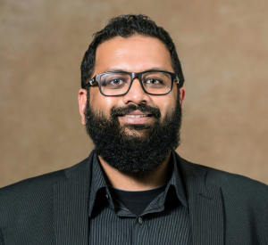 A man with short black hair and a beard wearing black glasses, a black suit jacket over a dark gray striped collared shirt.