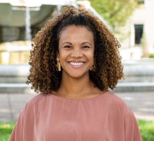 Woman with brown curly hair wearing a peach blouse and gold earrings.