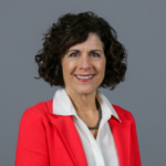 Woman with short, black curly hair wearing an orange blazer over a white collared shirt with a dark beaded necklace.