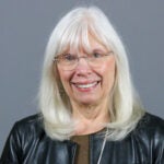 Woman with white hair wearing silver glasses, a black leather jacket and a brown top.