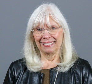 Woman with white hair wearing silver glasses, a black leather jacket and a brown top.