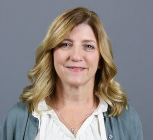 Woman with blonde hair wearing a gray cardigan over a white blouse and a silver necklace.