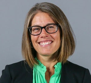 Woman with short brown hair wearing a black blazer over a bright green blouse and black glasses.