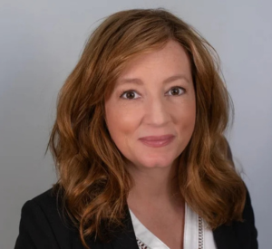 Woman with auburn hair wearing a black blazer over a white blouse.