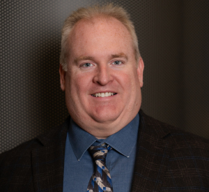 Man with short blonde hair wearing a black suit jacket over a dark green collared shirt with a striped tie.