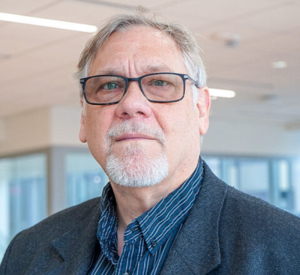 Man with short gray hair wearing a dark gray suit jacket over a dark blue striped collared shirt.