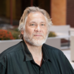 Man with short white hair and a white beard wearing a black collared shirt with vertical black stripes.