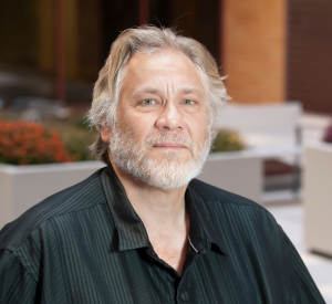 Man with short white hair and a white beard wearing a black collared shirt with vertical black stripes.