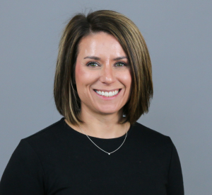 Woman with short brown hair with highlights wearing a long sleeve black top and a gold necklace.