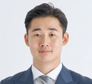 Man with short black hair wearing a blue suit jacket over a white collared shirt with a gray striped tie.