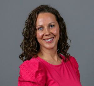 Woman with brown curly hair wearing a pink blouse with puffy sleeves.