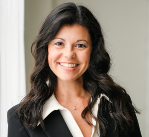 Woman with brown curled hair wearing a black blazer over a white collared shirt and a silver necklace.