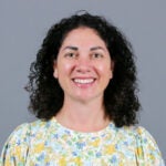 Woman with dark brown curly hair wearing a floral blouse with yellow, pink, blue and green flowers.