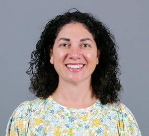 Woman with dark brown curly hair wearing a floral blouse with yellow, pink, blue and green flowers.