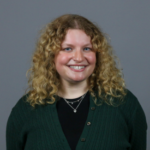 Woman with dark blonde curly hair wearing a green sweater over a black top and silver necklaces.