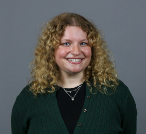 Woman with dark blonde curly hair wearing a green sweater over a black top and silver necklaces.