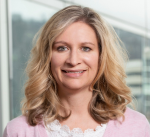 Woman with blonde curled hair wearing a pink cardigan over a white blouse with floral detailing.