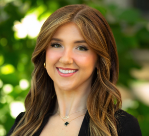 Woman with auburn hair wearing a black blazer and a gold and black necklace.