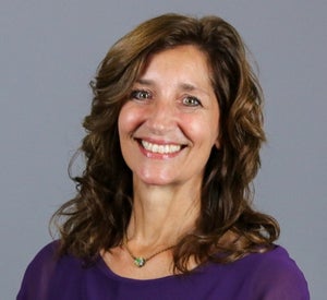 Woman with brown curly hair wearing a purple blouse and a necklace with a green pendant.